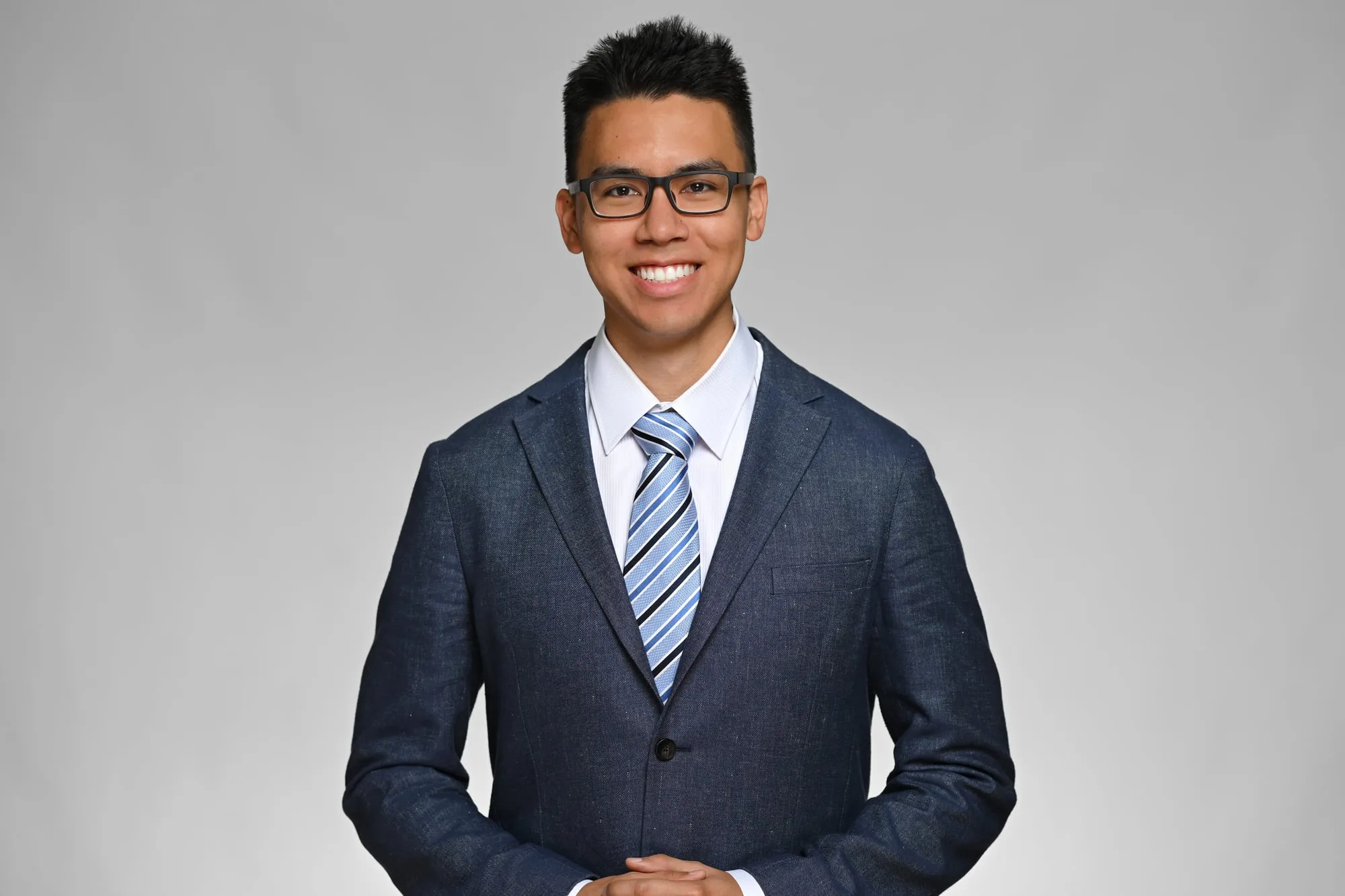 ERAS-ready medical residency headshot: navy suit, blue tie, and glasses, grey background