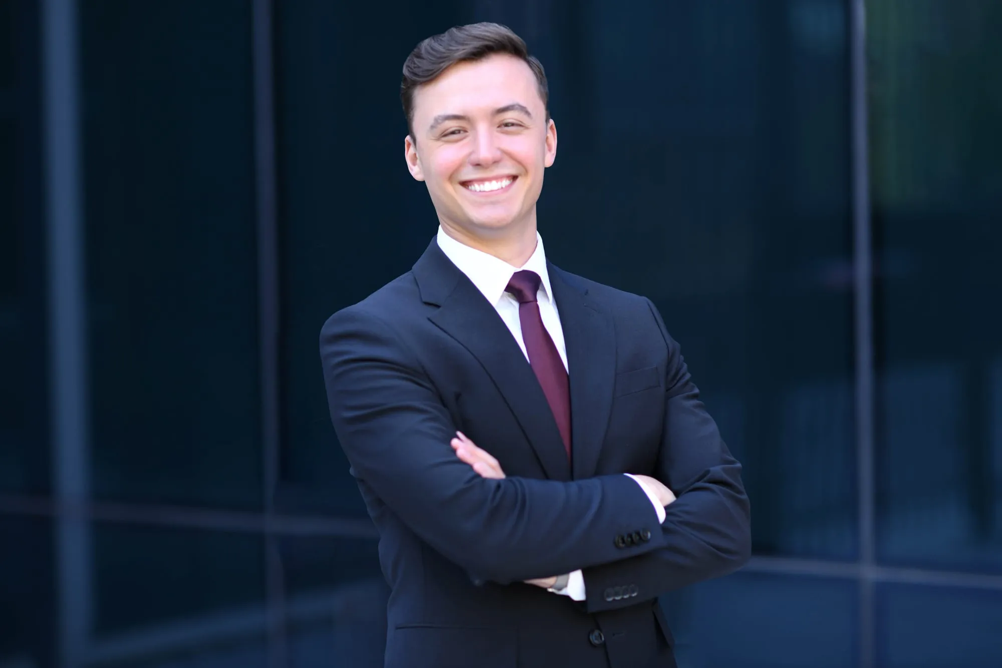 On-location headshot in Denver: navy suit and burgundy tie, arms crossed, glass building behind