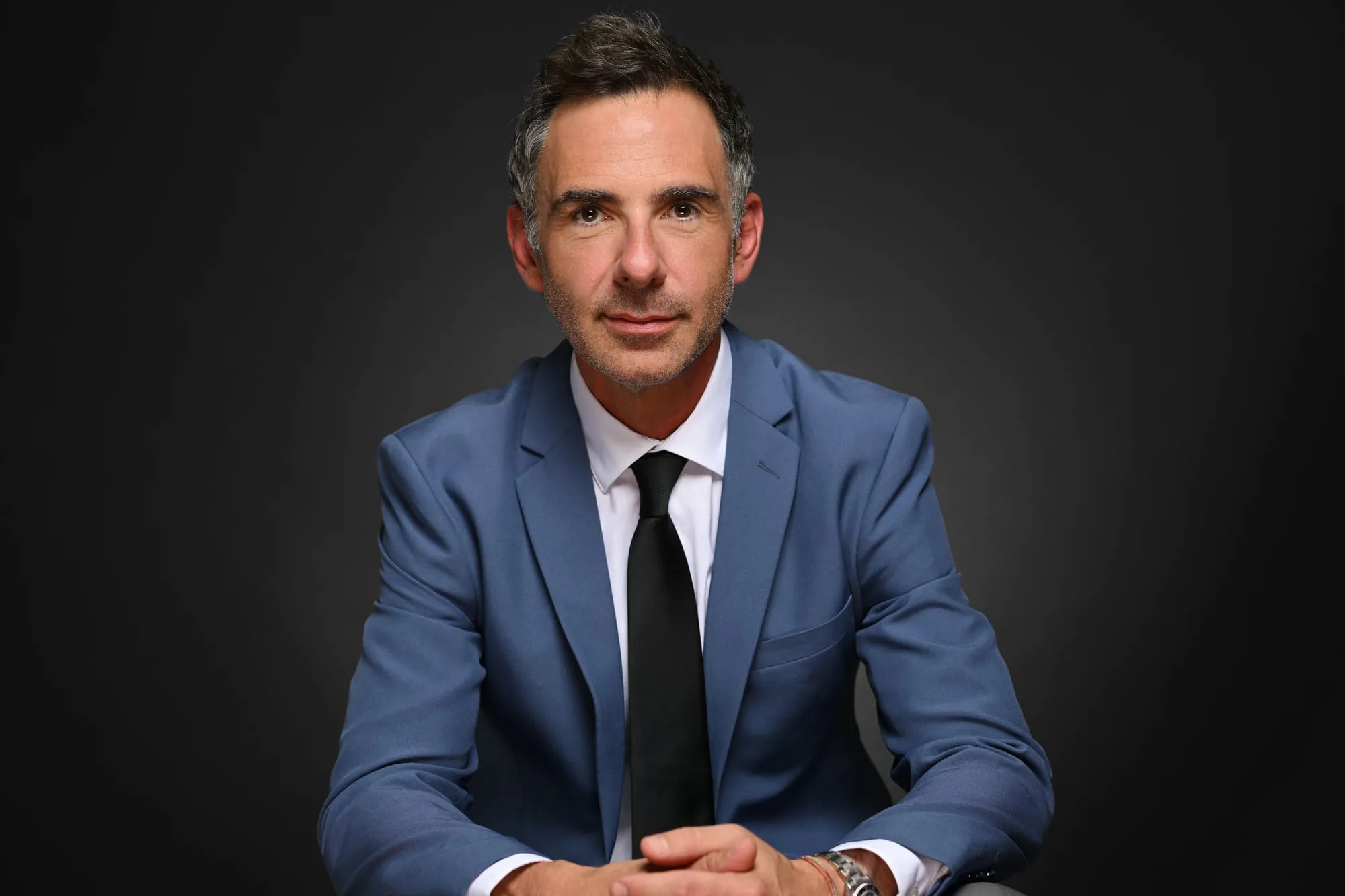 Studio headshot on a black background: blue suit and black tie, seated