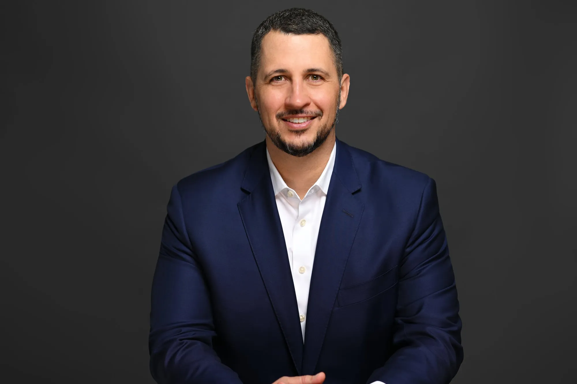 Studio headshot on a black background: navy suit and white shirt, seated