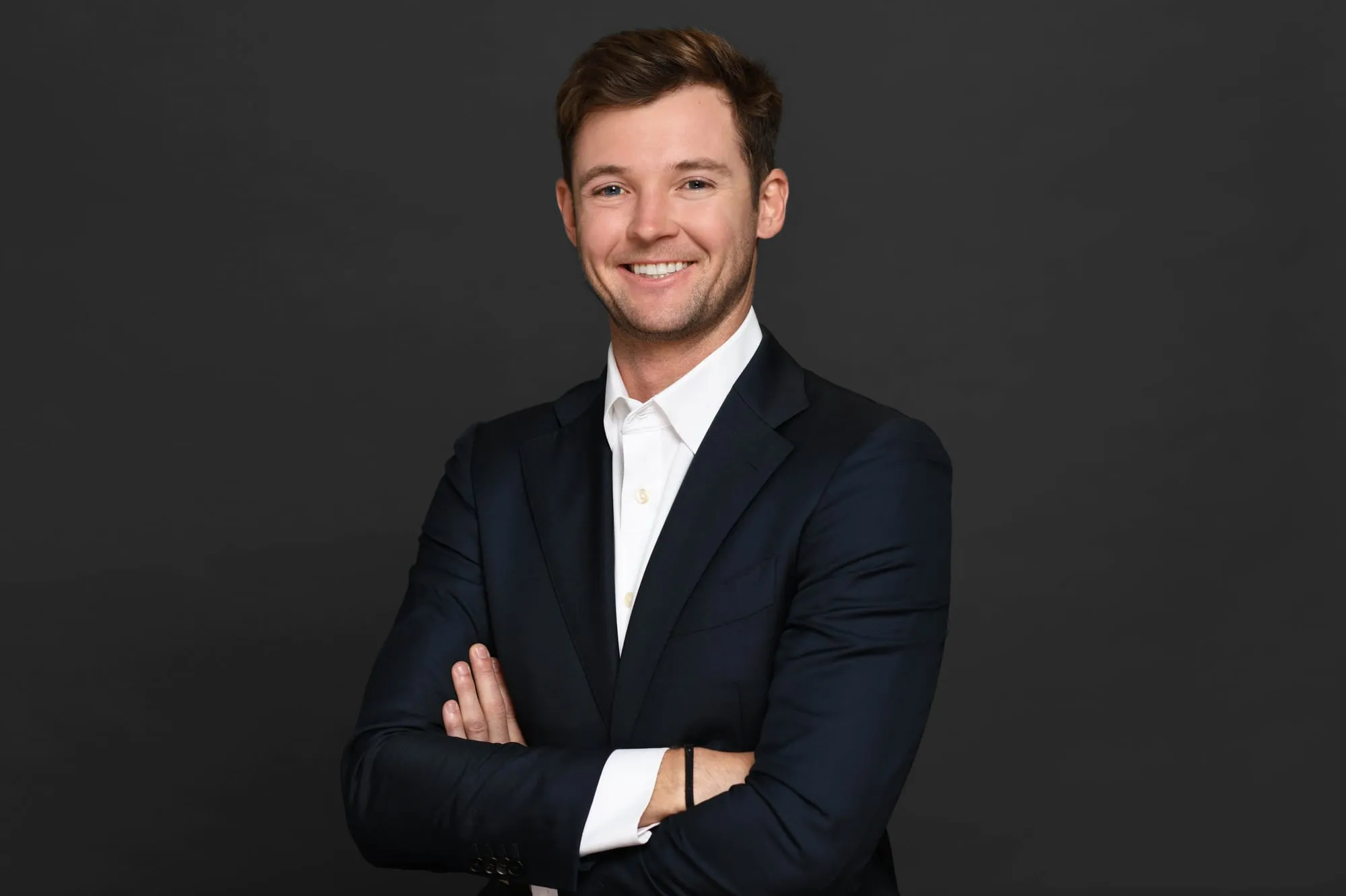 Studio headshot on a black background: navy suit, arms crossed