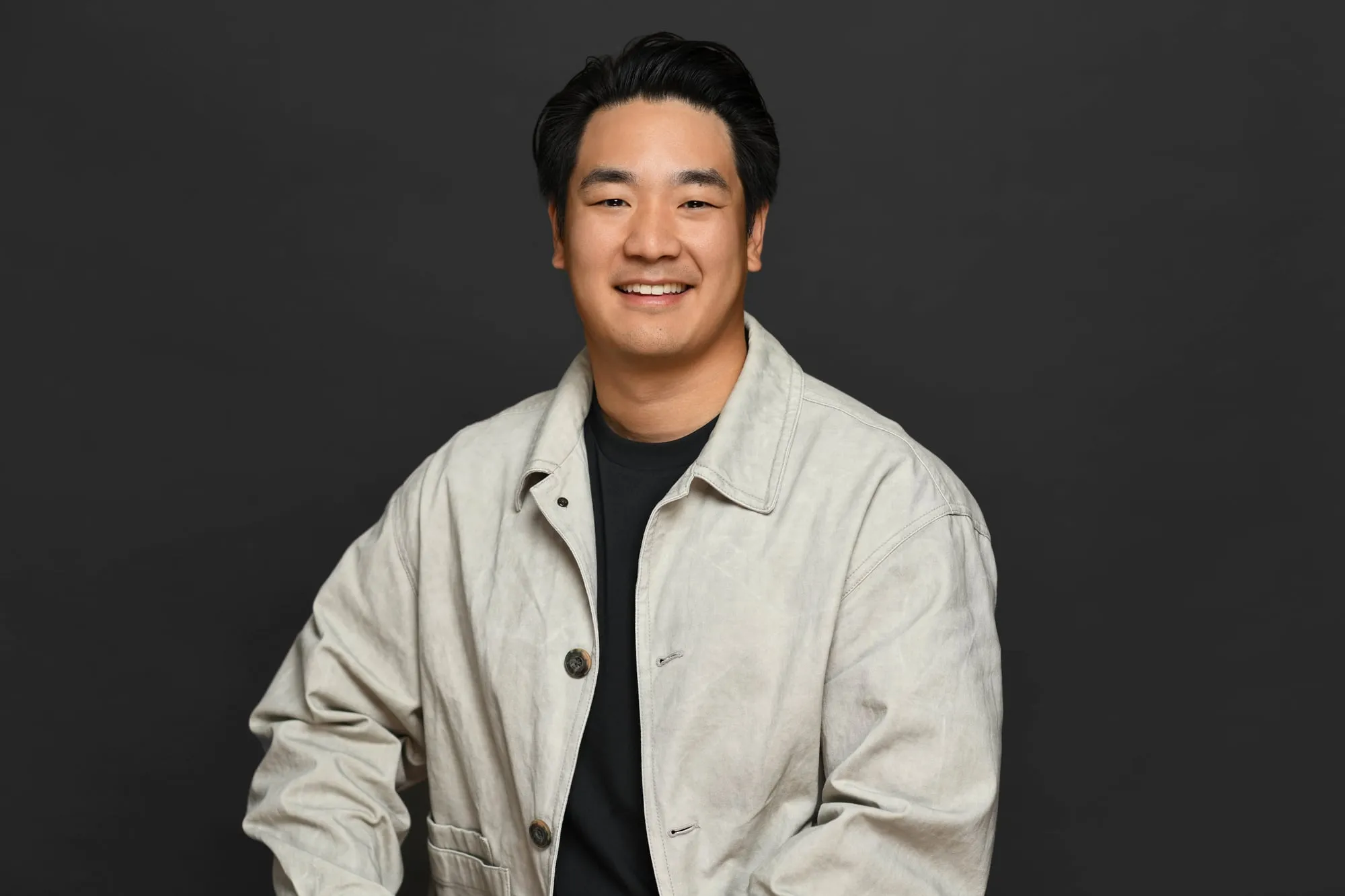 Studio headshot on a black background: light utility jacket over a black tee
