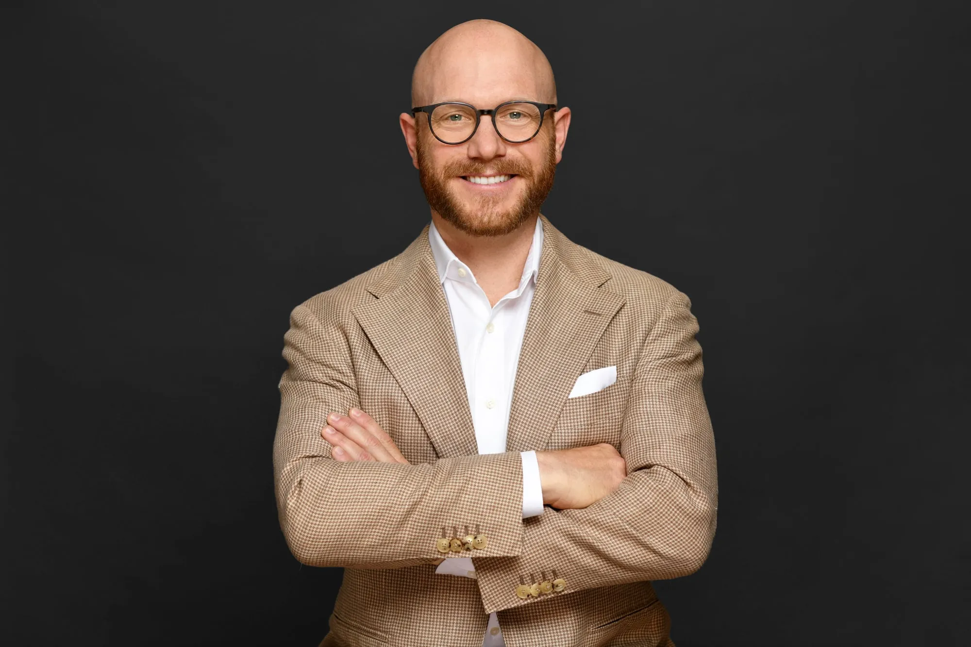 Studio headshot on a black background: tan double-breasted blazer and glasses, arms crossed