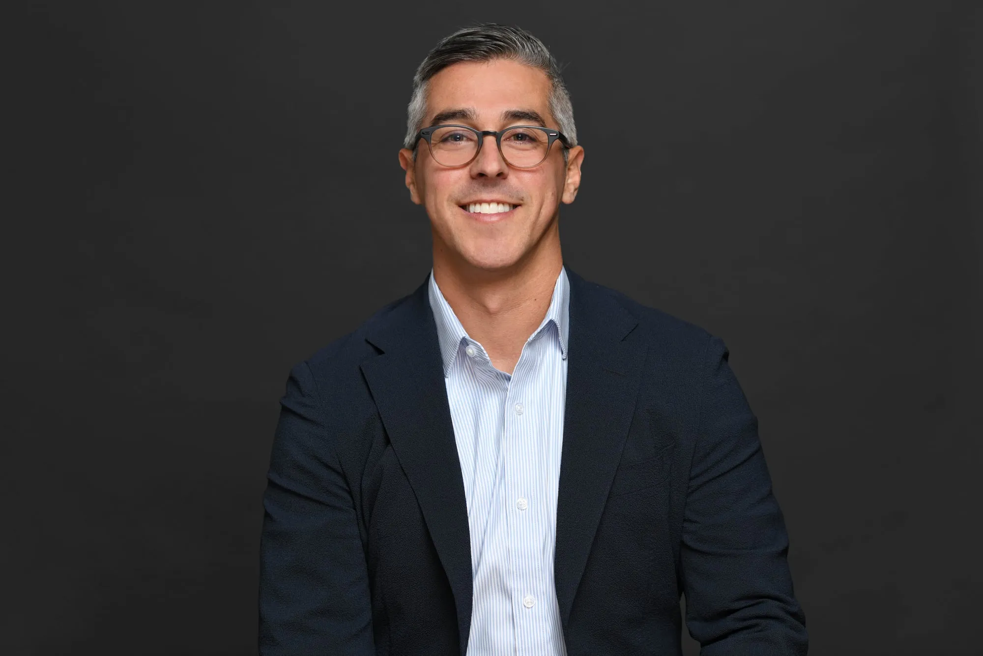 Studio headshot on a black background: navy blazer, light blue shirt, and glasses