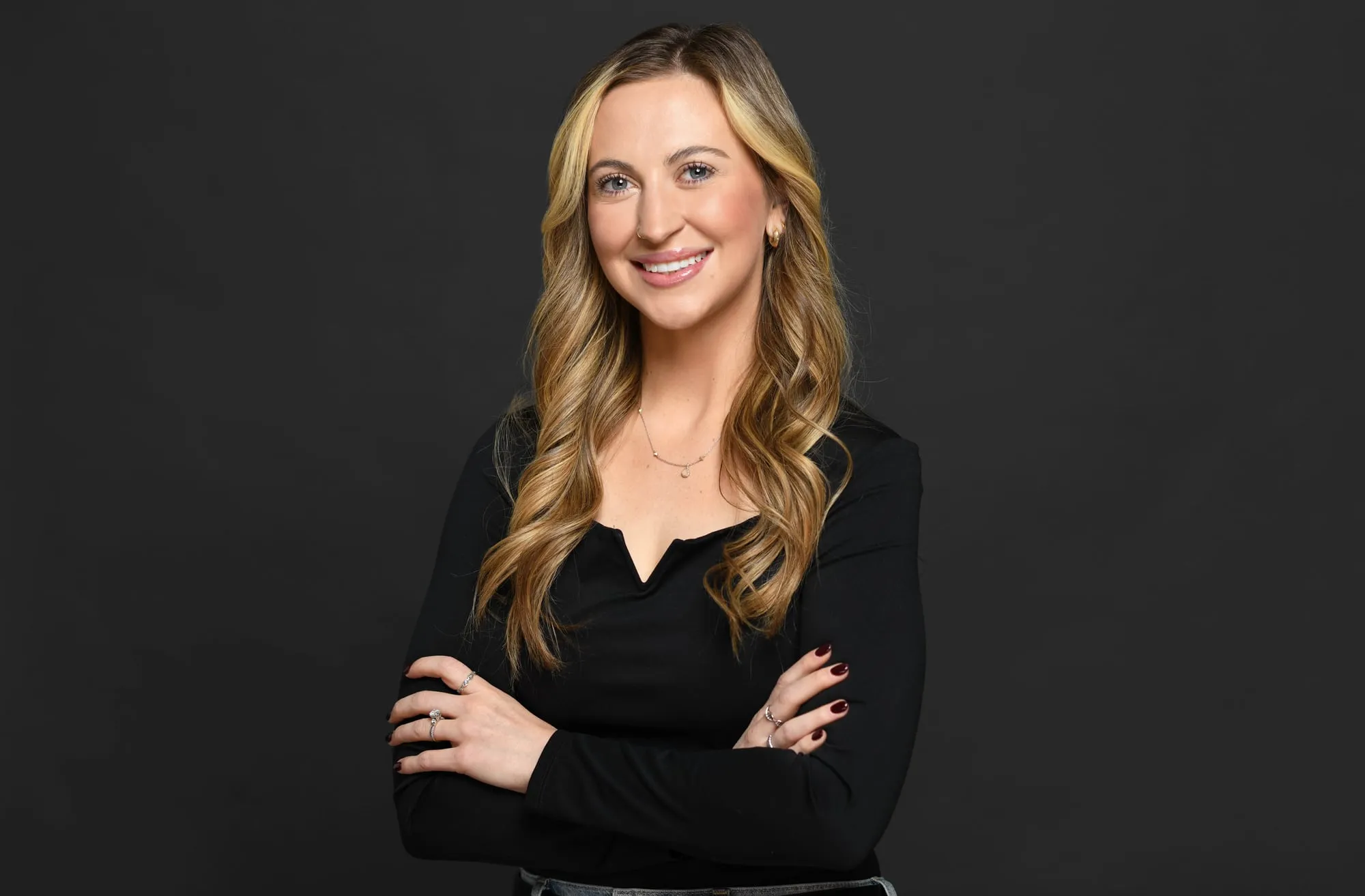Studio headshot on a black background: black top and long blonde hair, arms crossed