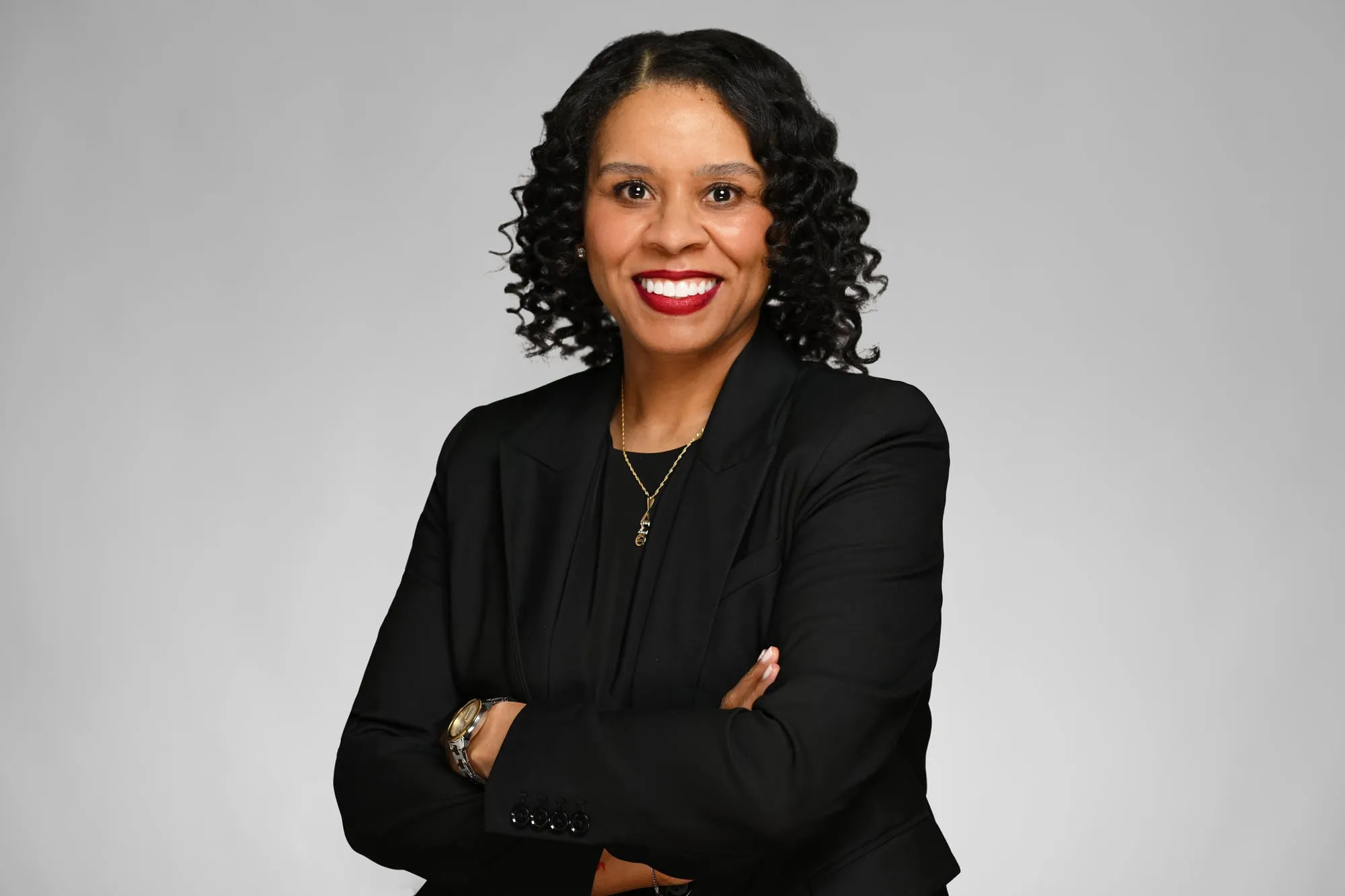 Studio headshot on a grey background: black blazer and curly hair, arms crossed