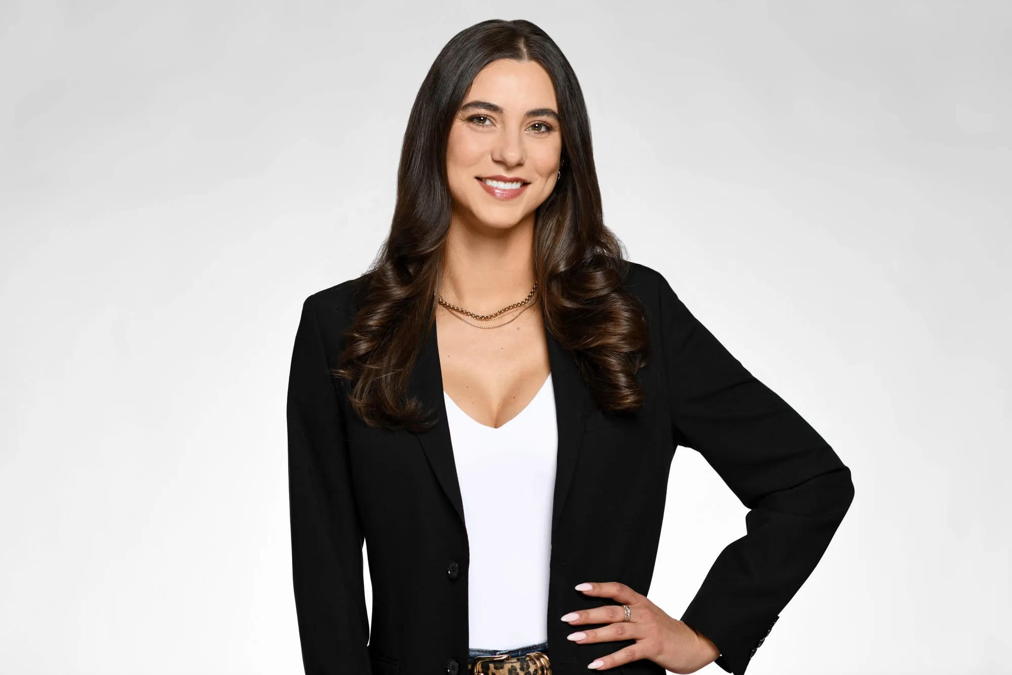 Studio headshot on a grey background: black blazer over a white top, hand on hip
