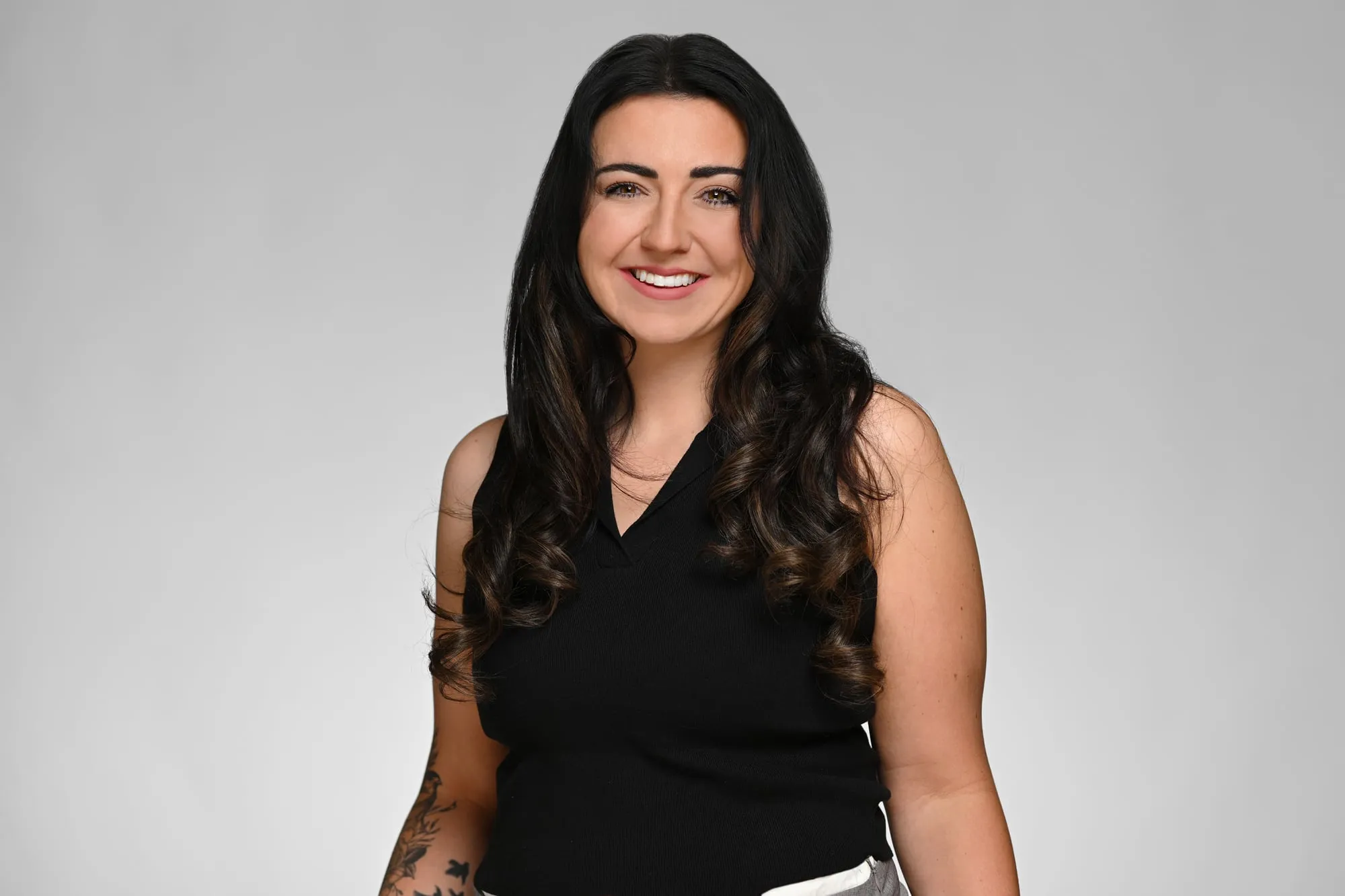 Studio headshot on a grey background: black sleeveless top and long dark hair