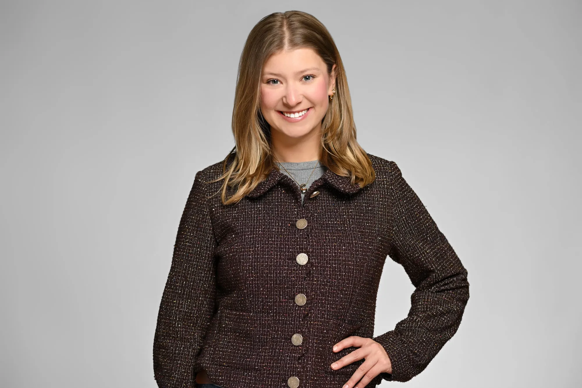 Studio headshot on a grey background: textured brown jacket, hand on hip