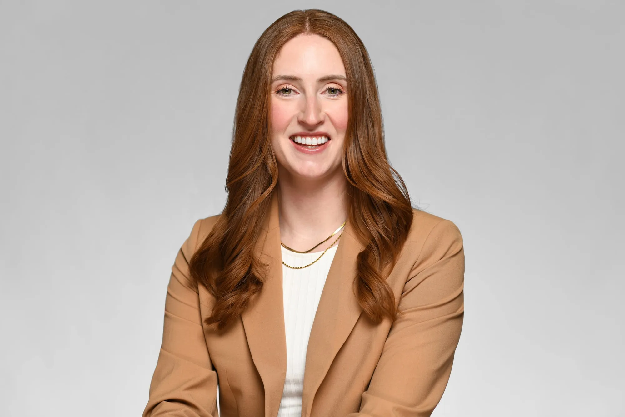 Studio headshot on a grey background: camel blazer and long auburn hair