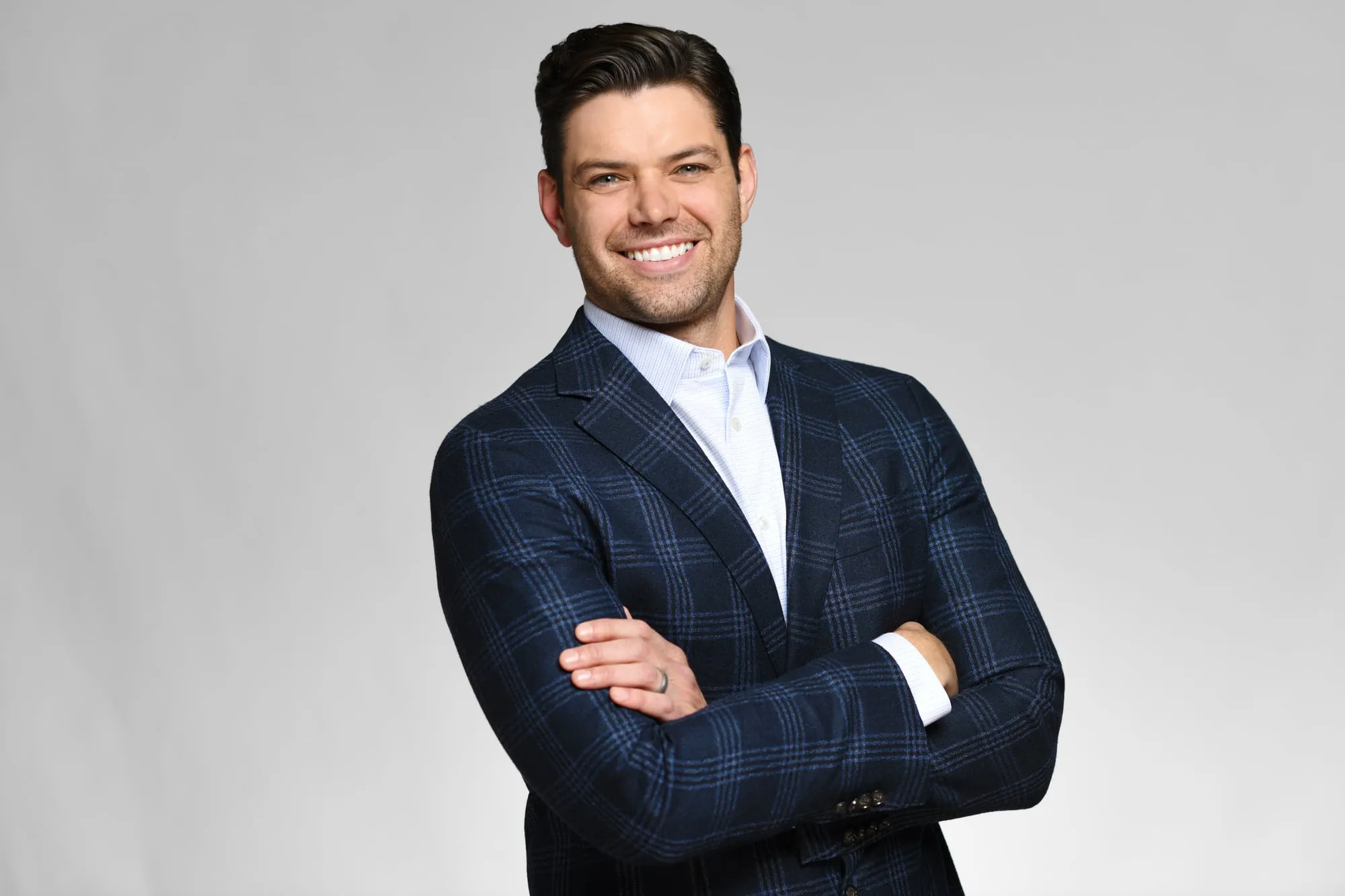 Studio headshot on a grey background: navy check blazer, arms crossed