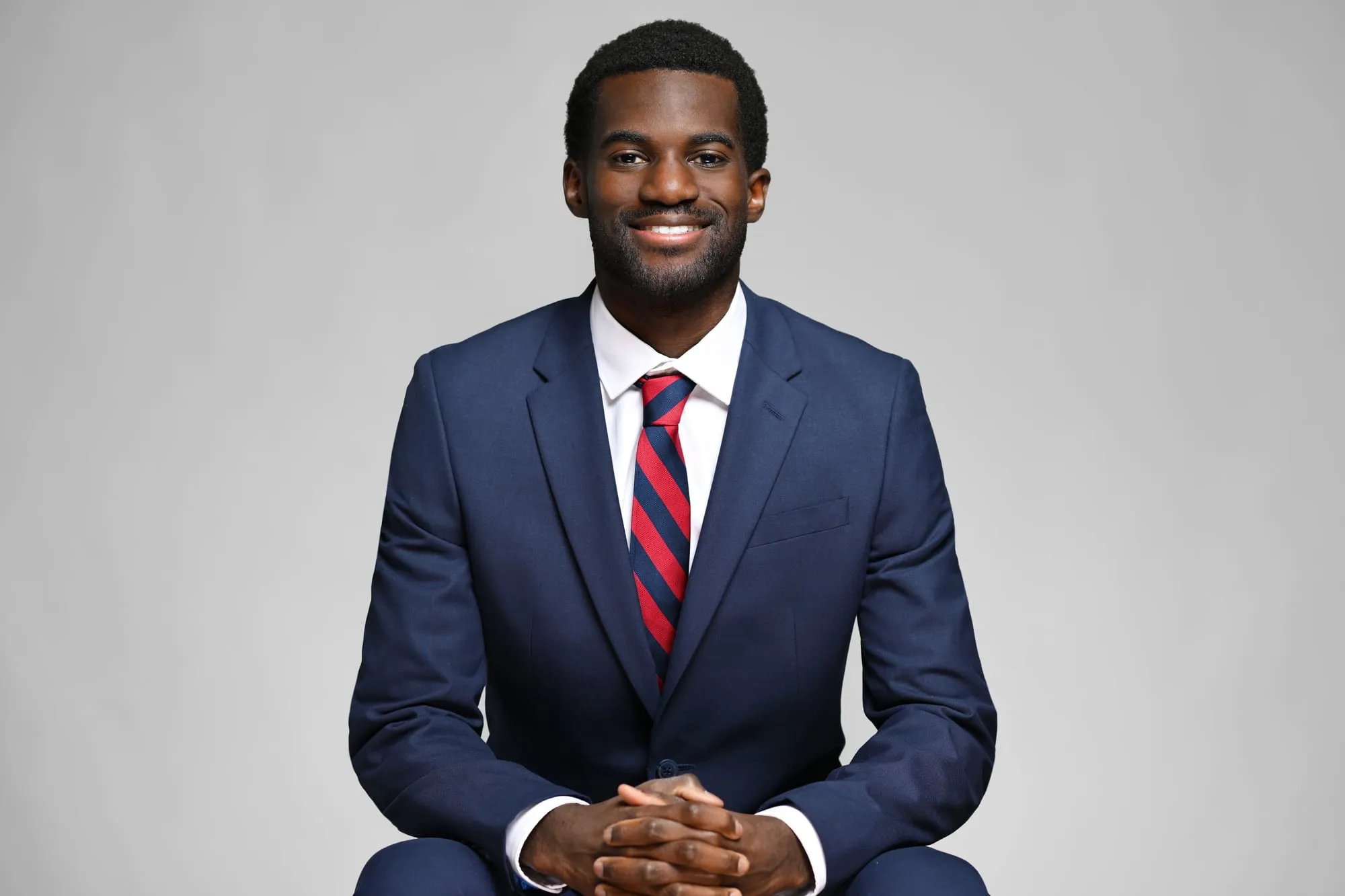 Studio headshot on a grey background: navy suit and striped red tie, seated