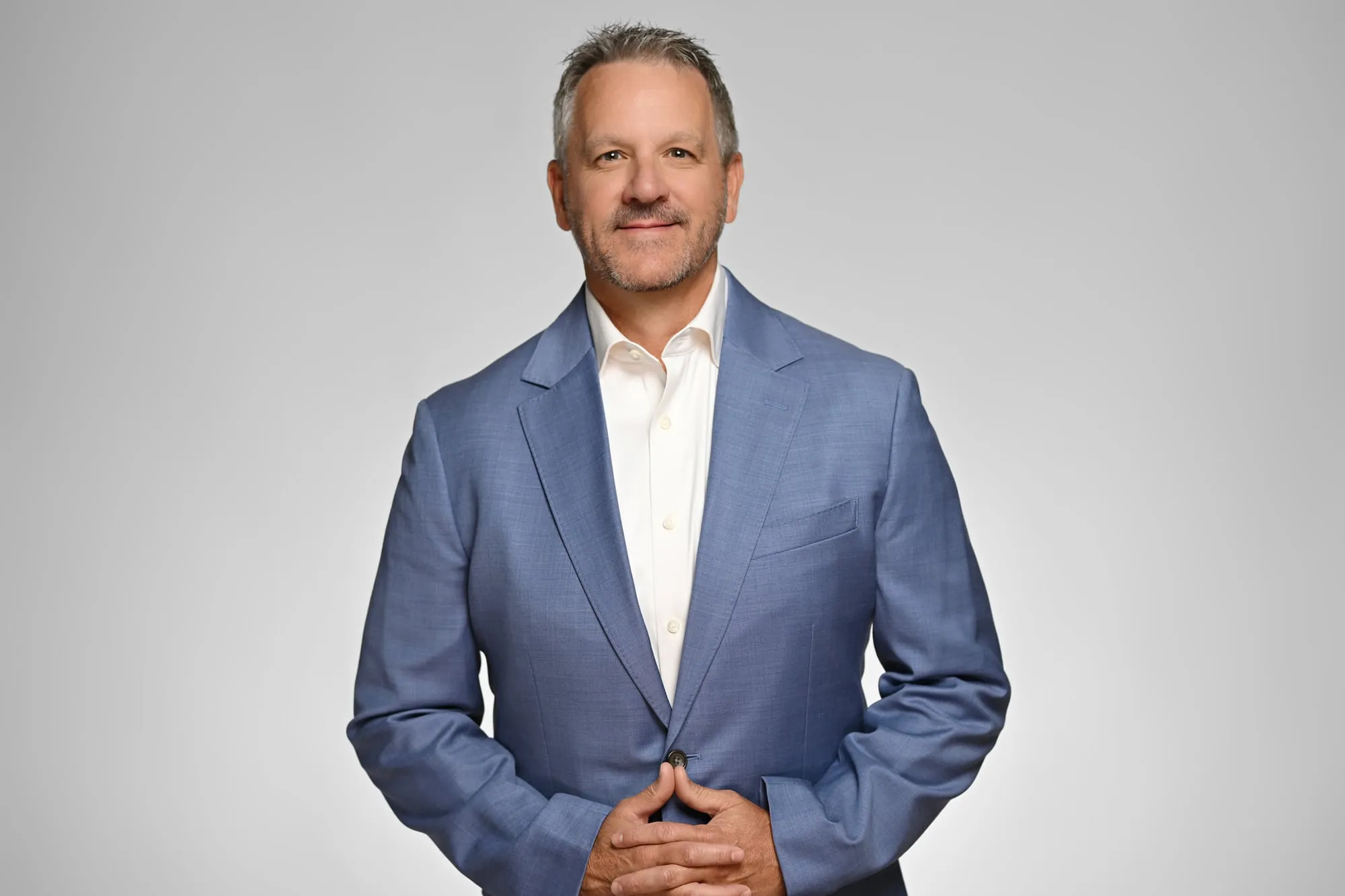 Studio headshot on a grey background: light blue blazer, seated