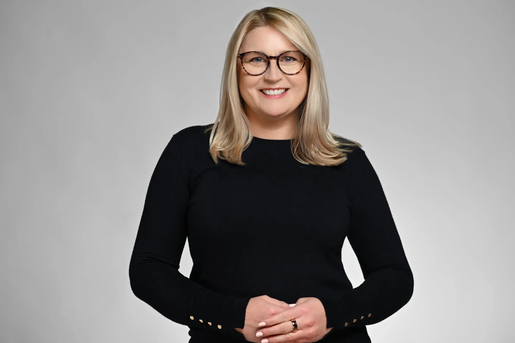Studio headshot on a grey background: black sweater and glasses