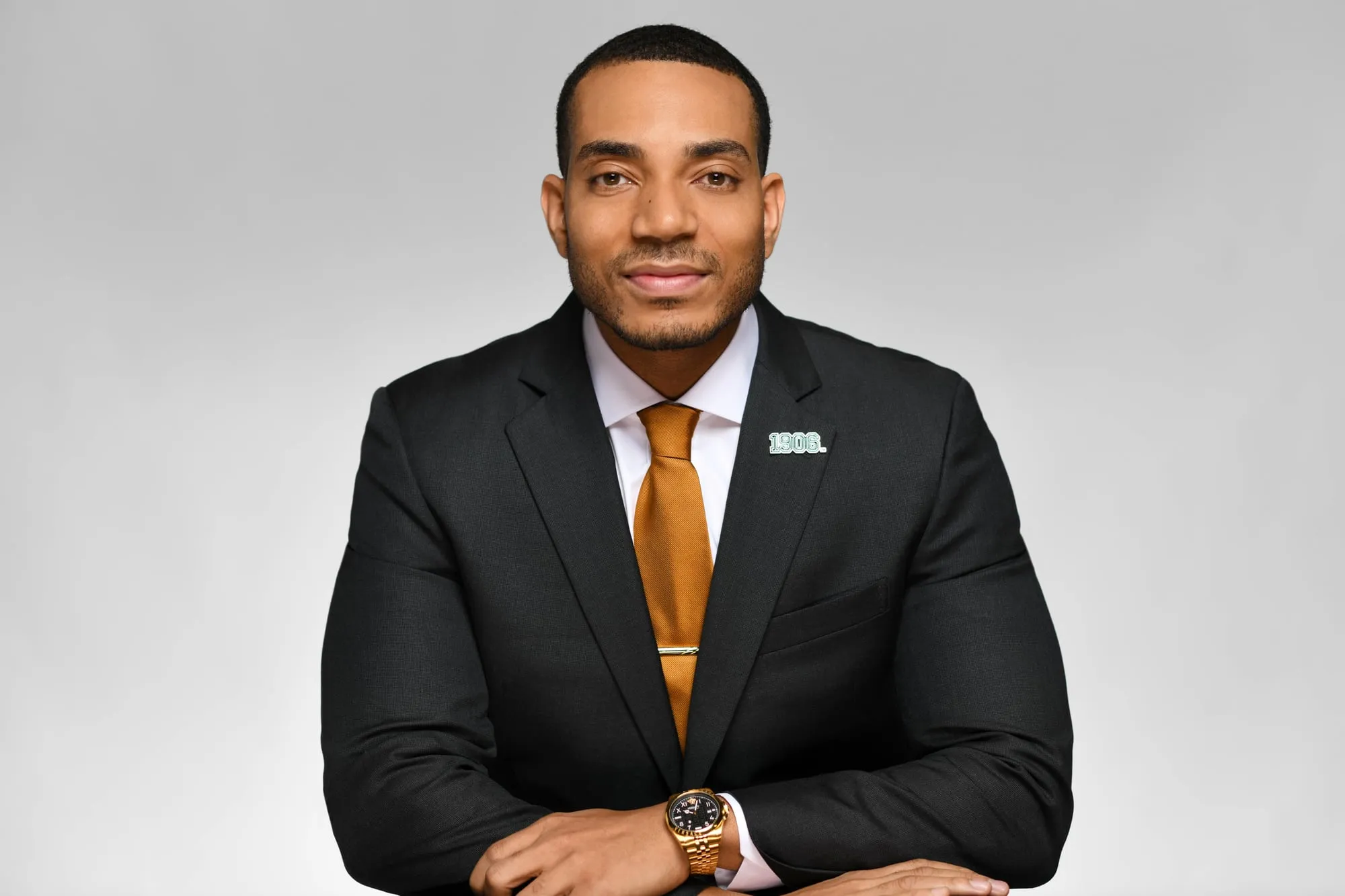 Studio headshot on a grey background: black suit and gold tie, seated