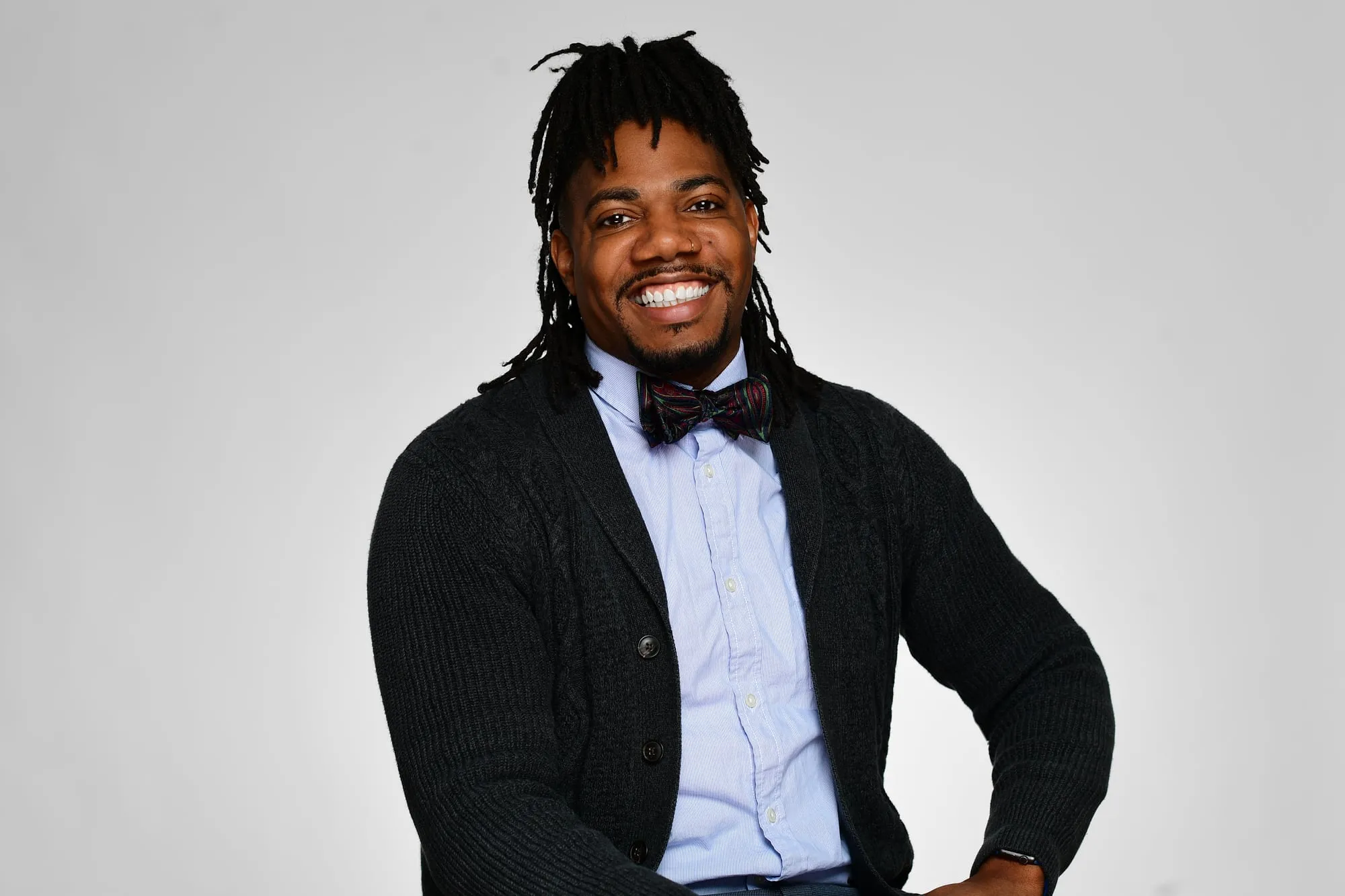 Studio headshot on a grey background: dark cardigan and bow tie, seated