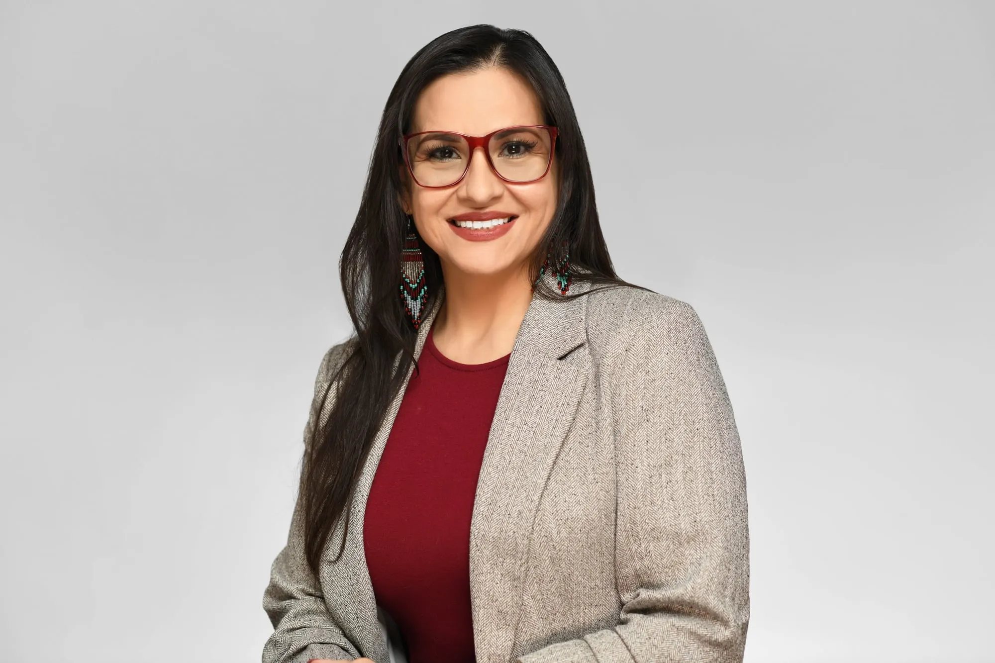 Studio headshot on a grey background: tan blazer over a red top, glasses