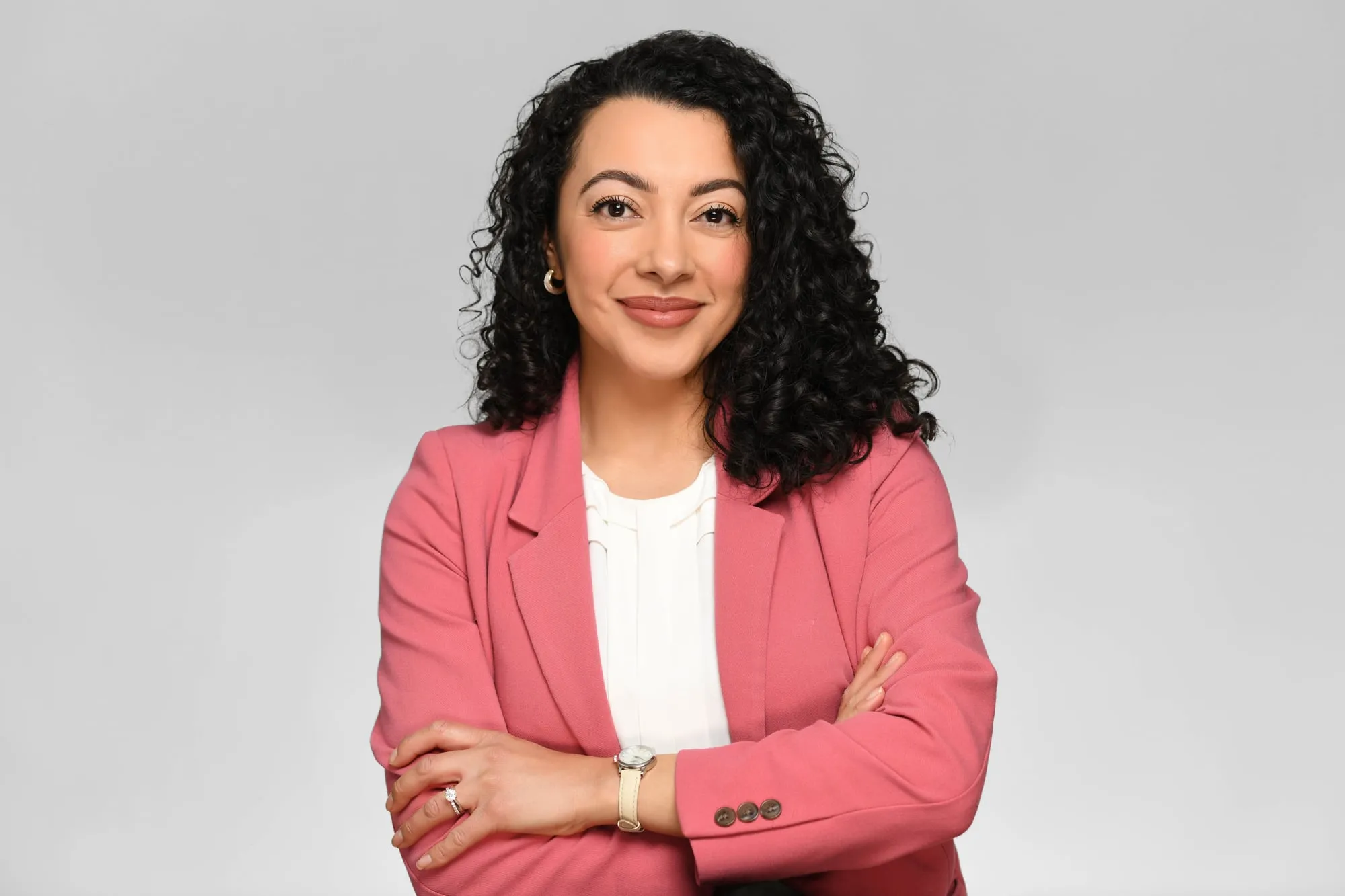 Studio headshot on a grey background: coral blazer and curly hair, arms crossed