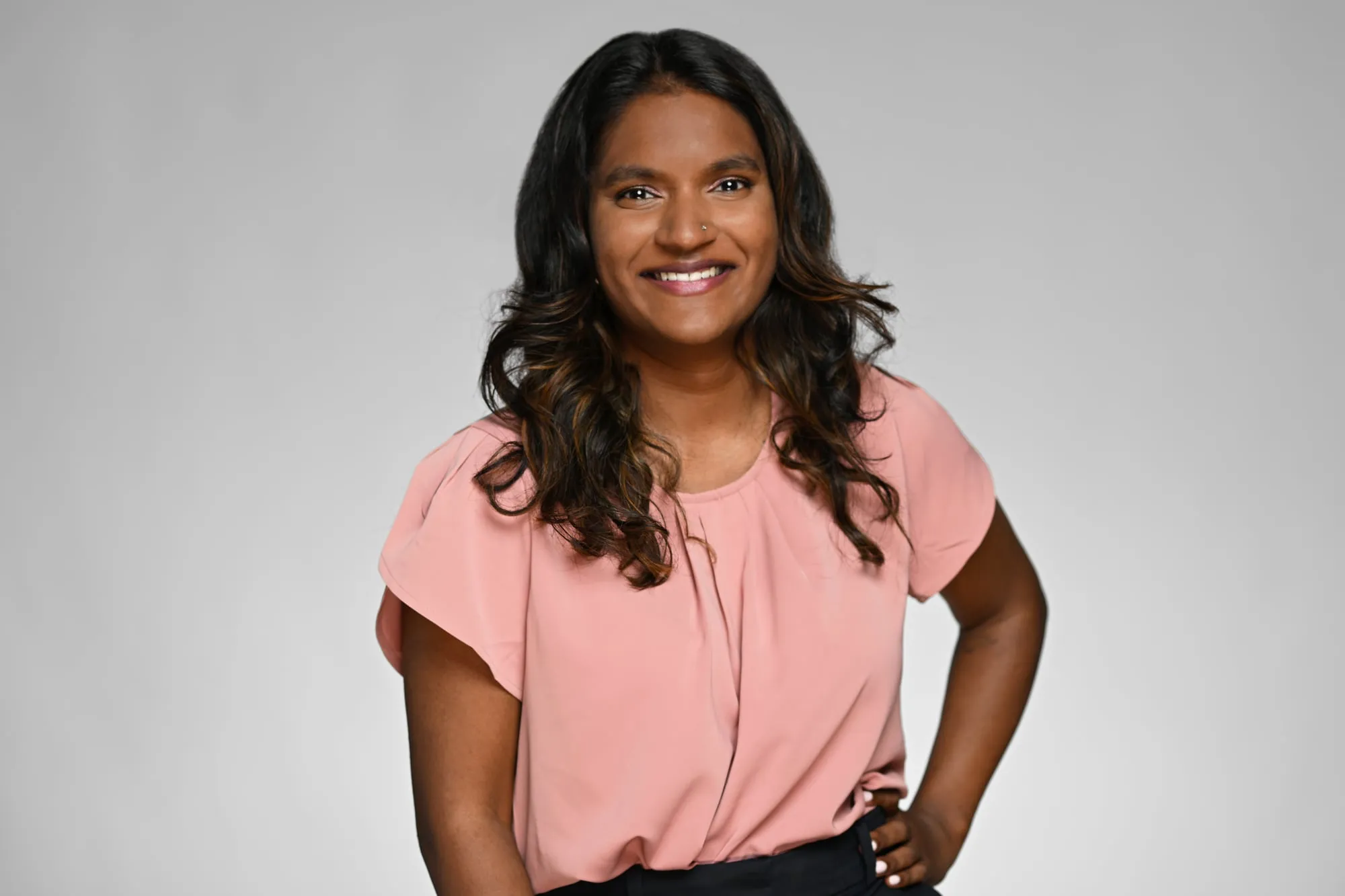Studio headshot on a grey background: pink blouse, hand on hip