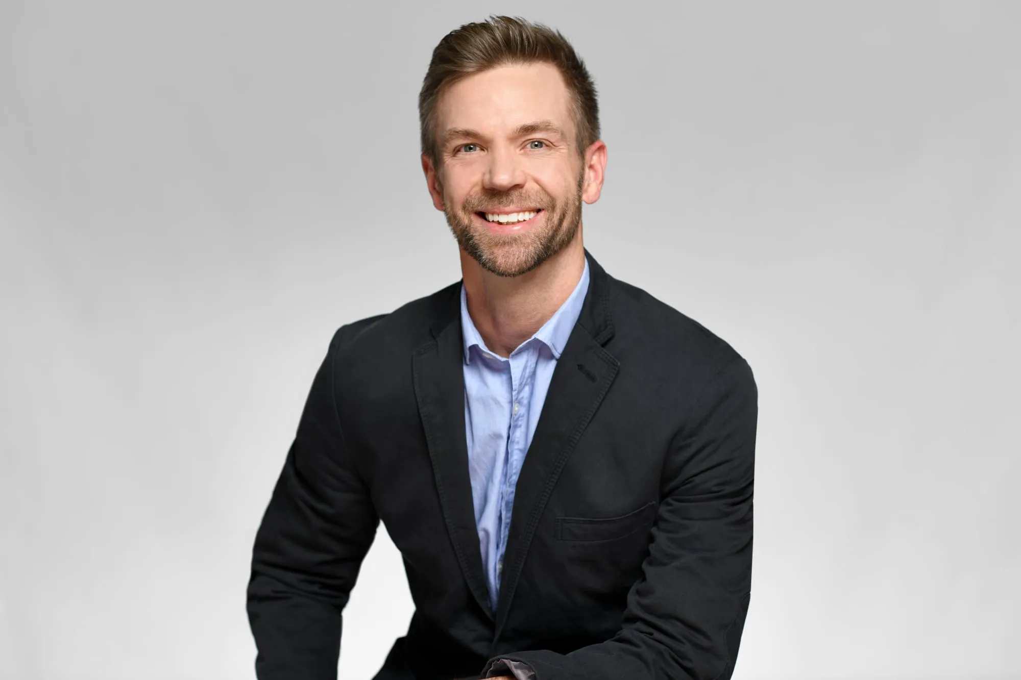 Studio headshot on a grey background: black blazer and light blue shirt