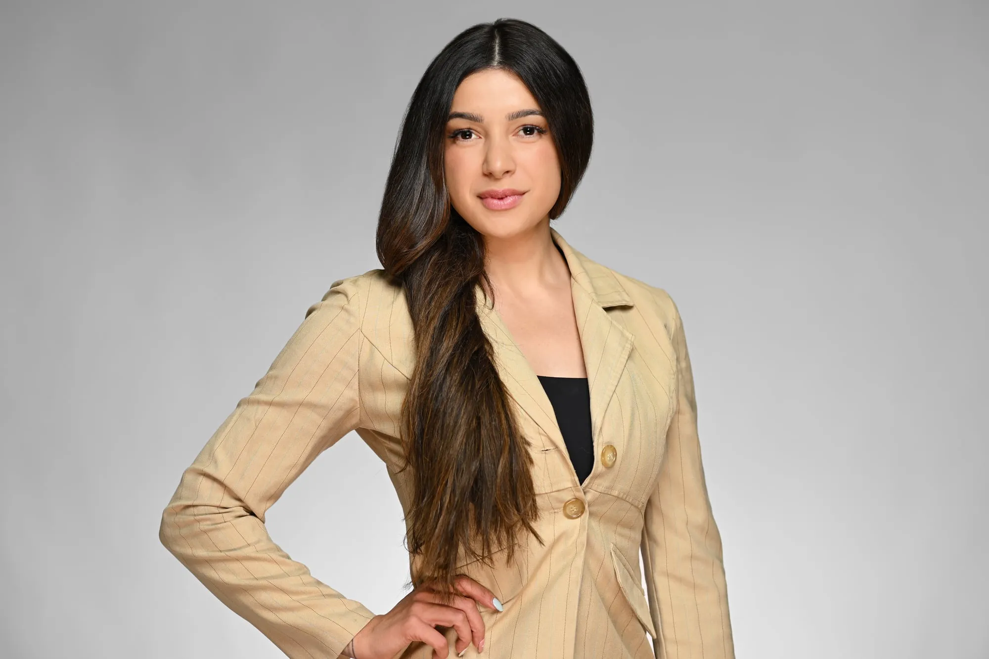 Studio headshot on a grey background: tan blazer and long hair, hand on hip