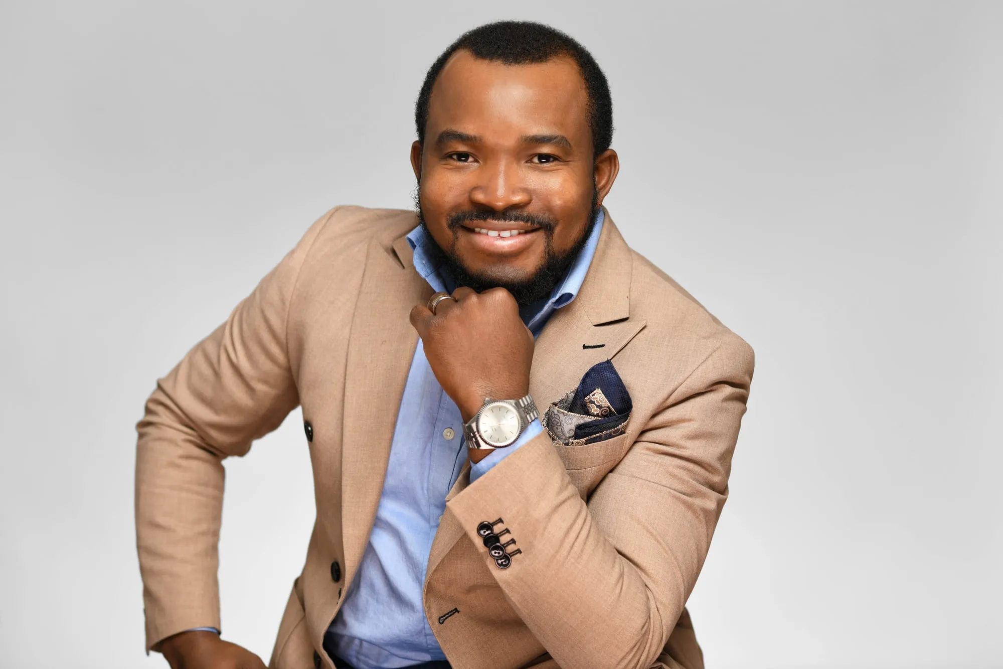 Studio headshot on a grey background: tan blazer, hand resting near the chin