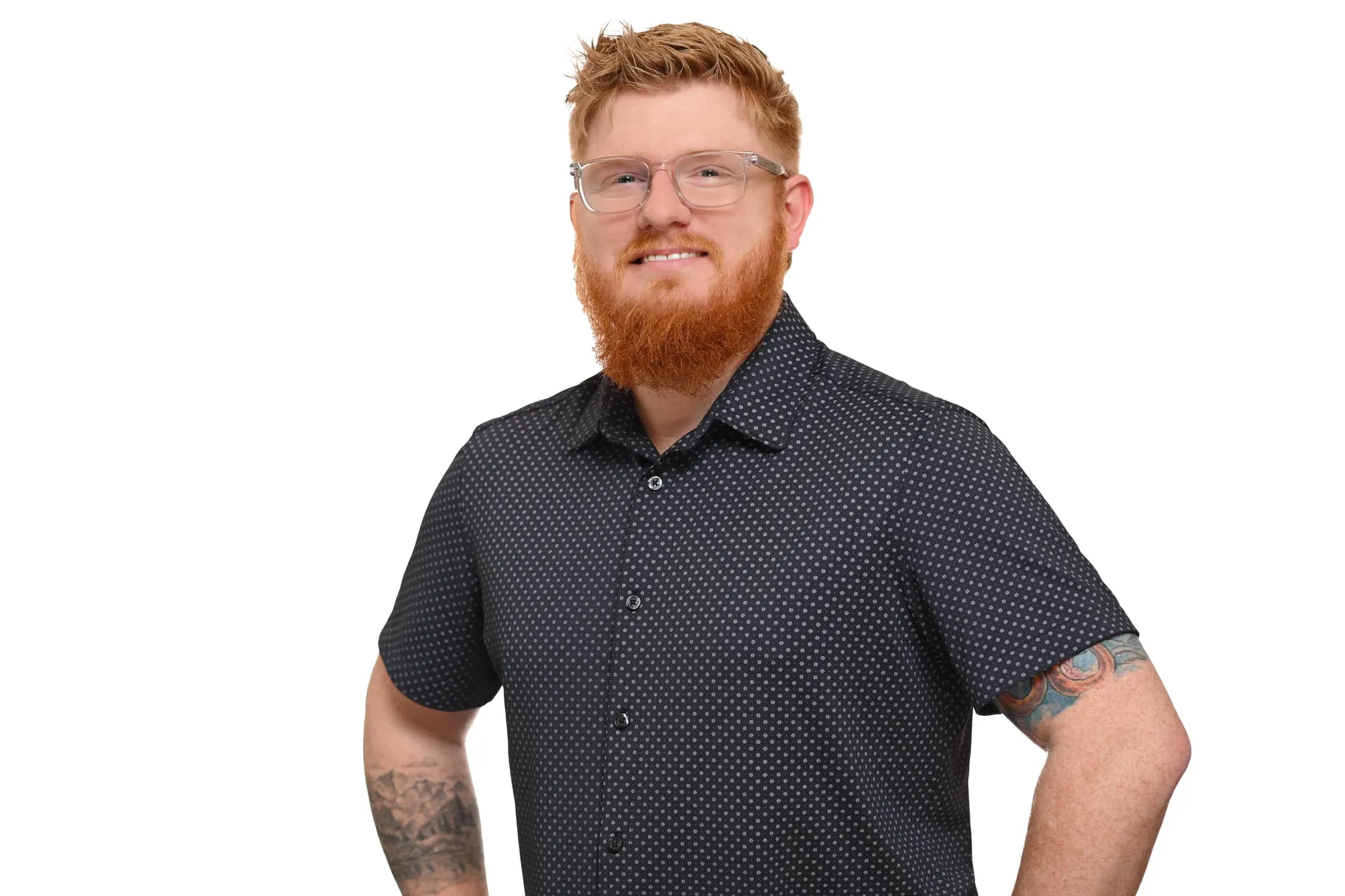 Studio headshot on a white background: patterned short-sleeve shirt, glasses, and a red beard