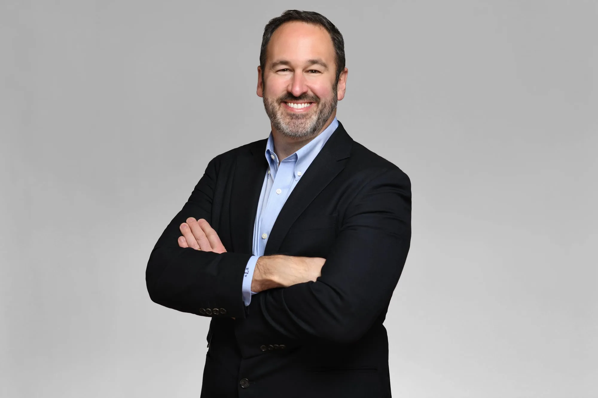 Corporate headshot for Tract in Denver on a light grey background: black blazer and light blue shirt, arms crossed